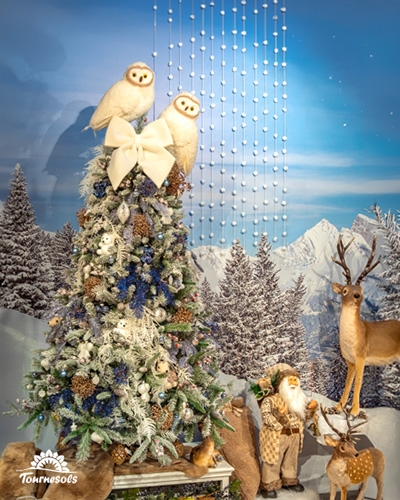 Sapin enneigé bleu et blanc coiffé de deux chouettes effraies, père Noël beige et cerfs devant un décor de montagne